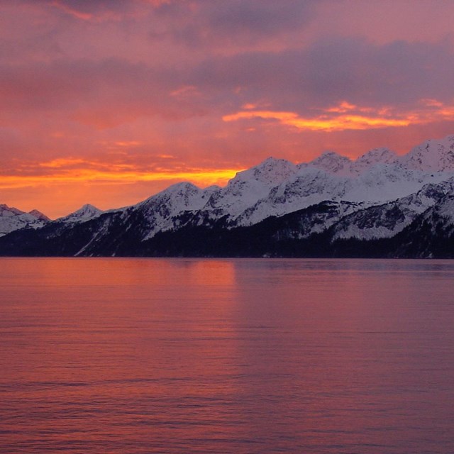 Rosy sunlight washes over snowy mountains in the distance; reflected off calm waters in foreground.