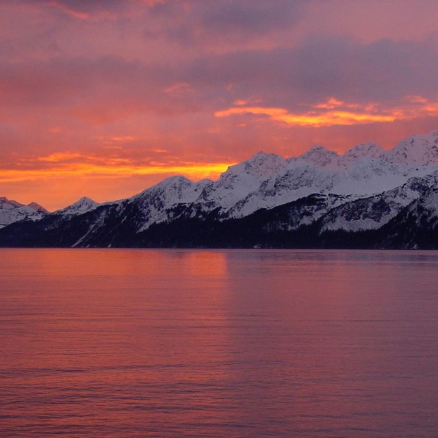 Rosy sunlight washes over snowy mountains in the distance; reflected off calm waters in foreground.