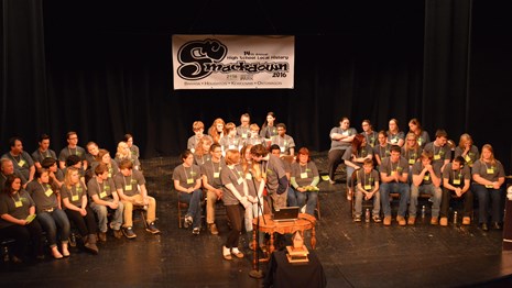 A group of students sits on a brightly lit stage.