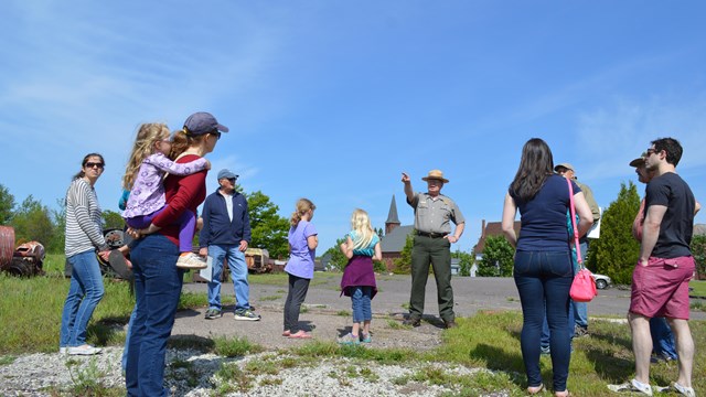 A park ranger points to something in the distance while a group of standing people look on.