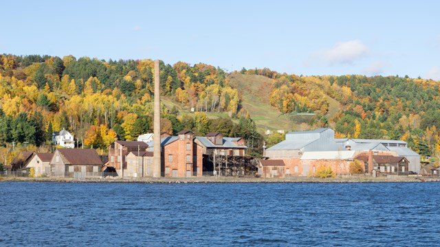 A large industrial complex at the foothills of a large hill situated along a waterway.
