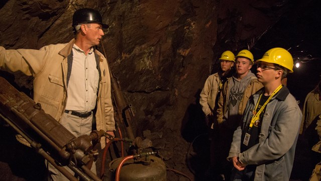 A tour guide with a hard hat talks to a group of youth with hard hats on about a drill used to mine.