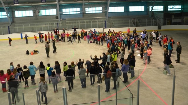 A large group of people stand in a half-circle on an indoor ice rink.