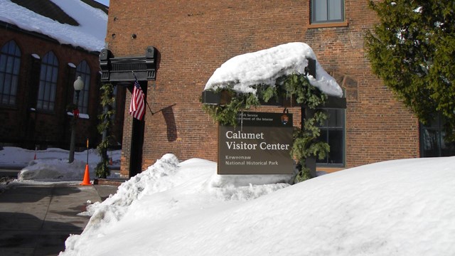 Sign reading "Calumet Visitor Center" in front or a tall brick building surrounded by snow.