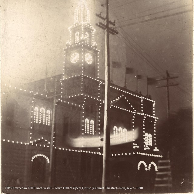 In this black and white historic image, Christmas lights illuminate a large stone building. 