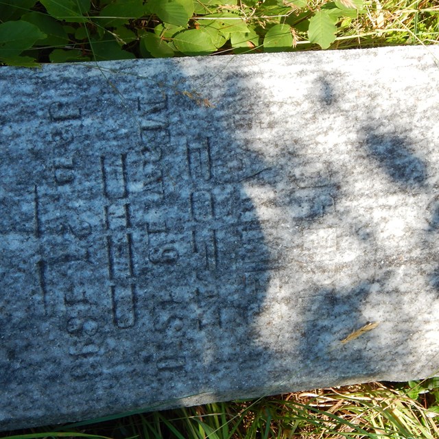 A grave marker on the ground shows a shadow made from a tree.
