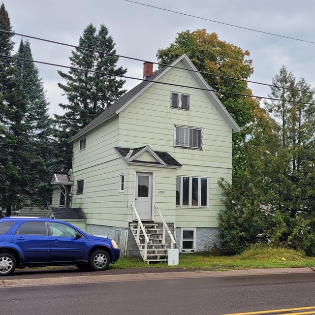 A two-story front-gabled frame house has a car parked alongside it. Stairs reach the elevated front 