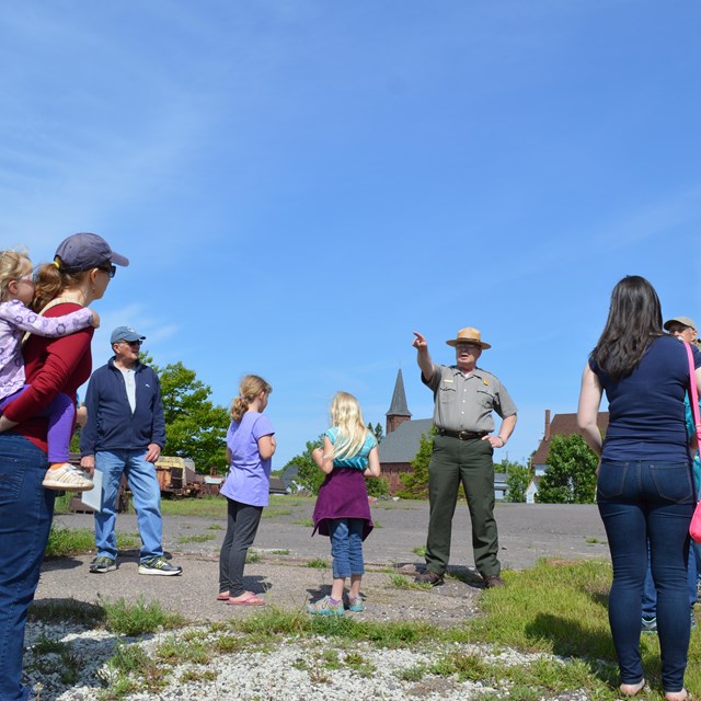A park ranger points to something in the distance while a group of standing people look on.