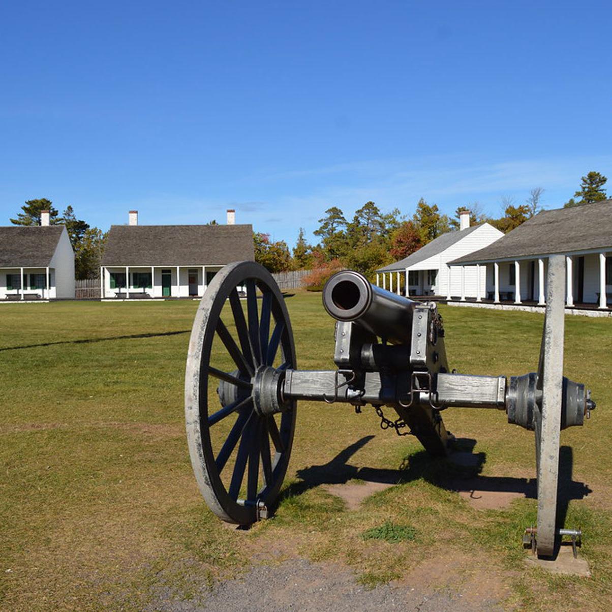 A historic cannon sits in a grassy field before many white wood one-story buildings with a blue sky.