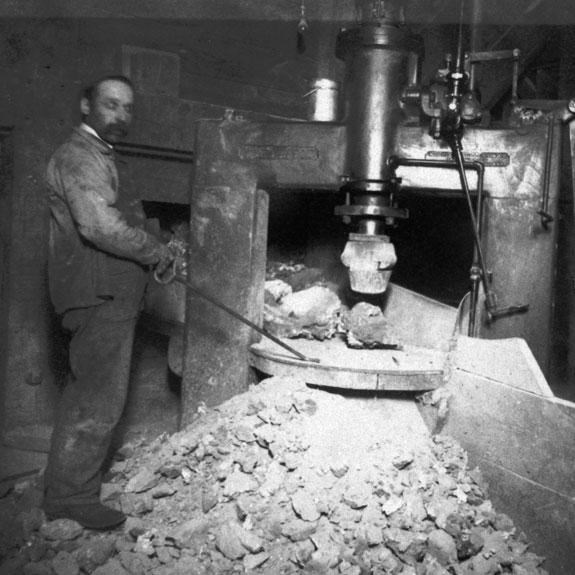 In this historic black and white image a man stands next to a rock crusher, another sits on right.