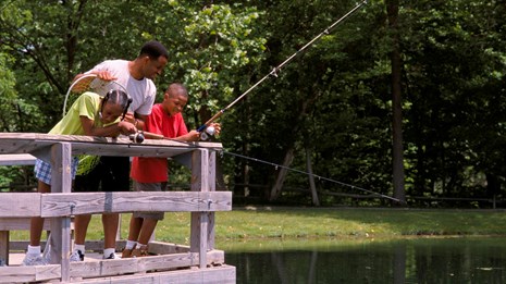 a family fishing from a pier with green trees and a grassy area in the background