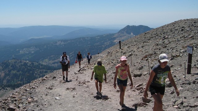 Six people hike up hill with a view of a valley in the background. 