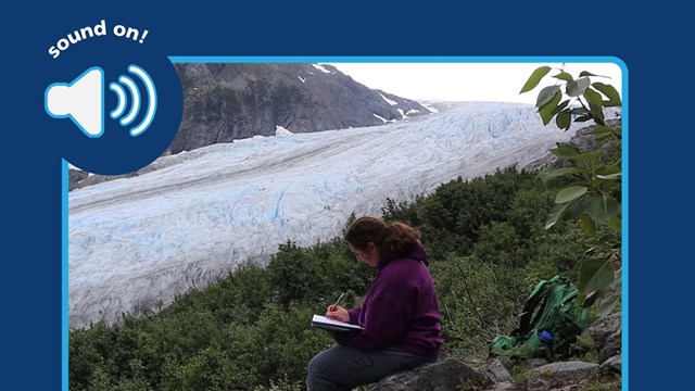 person sits by a large glacier, they have a notebook. Sound on icon.