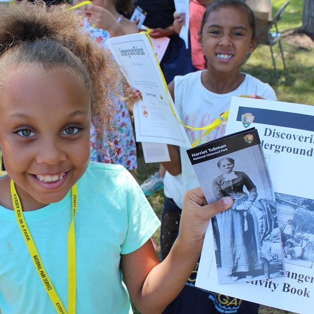 A young, smiling girl holds Junior Ranger booklet titled Discovering the Underground Railroad.