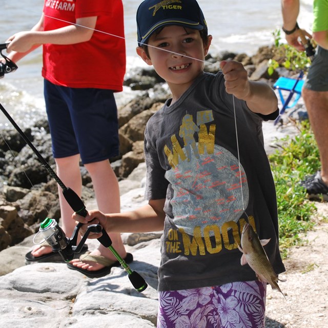 Boy stands along the shore with a fishing pole in hand.