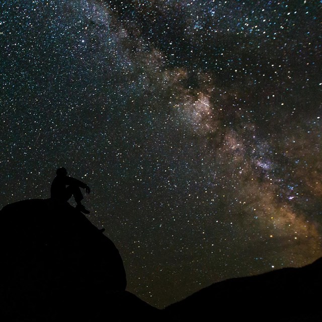 A person sits on a large rock gazing at a multitude of stars