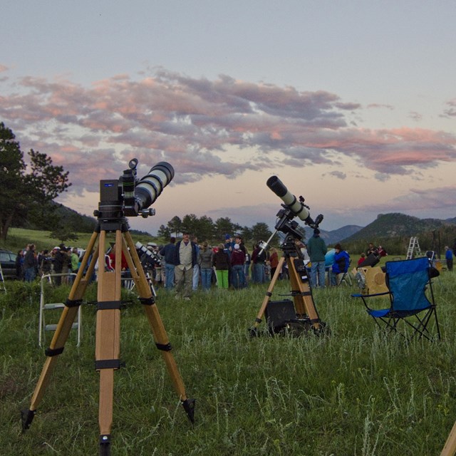 Yellow legged telescopes dot a grassy field at dusk. People are in the background.