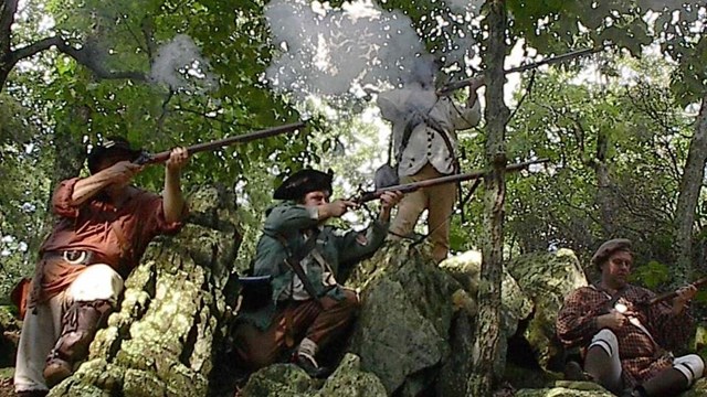 Living historians fire muskets while laying on rocks with trees in the background.