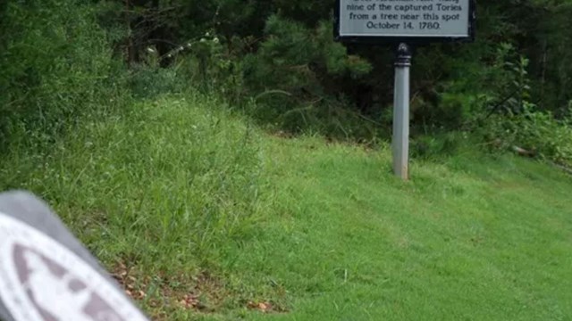 An image of a park sign and a historical marker along a road with trees in the background.