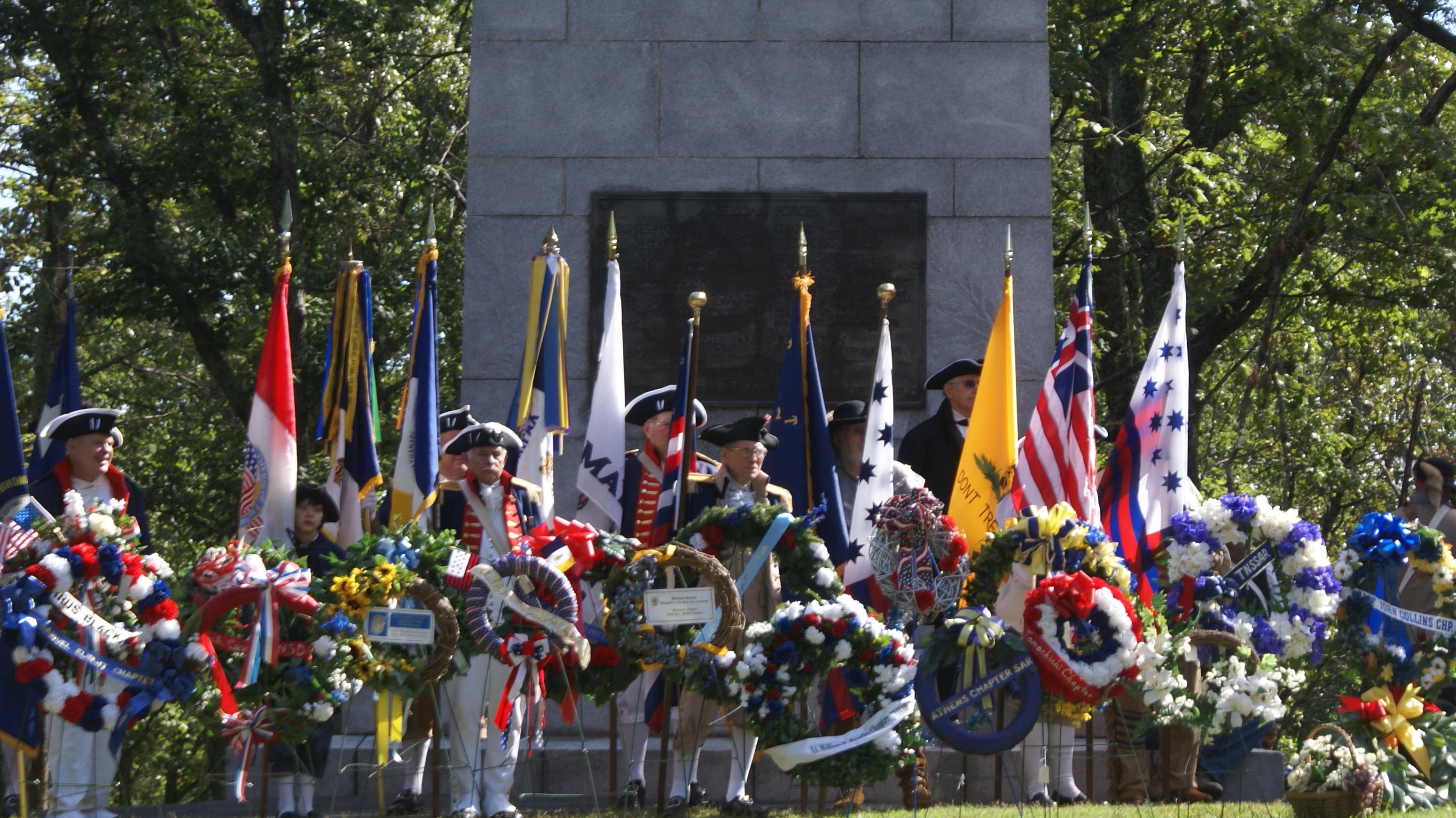 Living historians line up by wreaths with flowers by a large stone monument.
