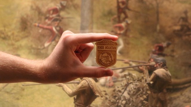 A hand holding a wooden badge that has a soldier firing a musket on it.