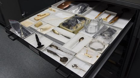 A shallow drawer pulled out to reveal a dozen artifacts in white boxes including spoons and glass.