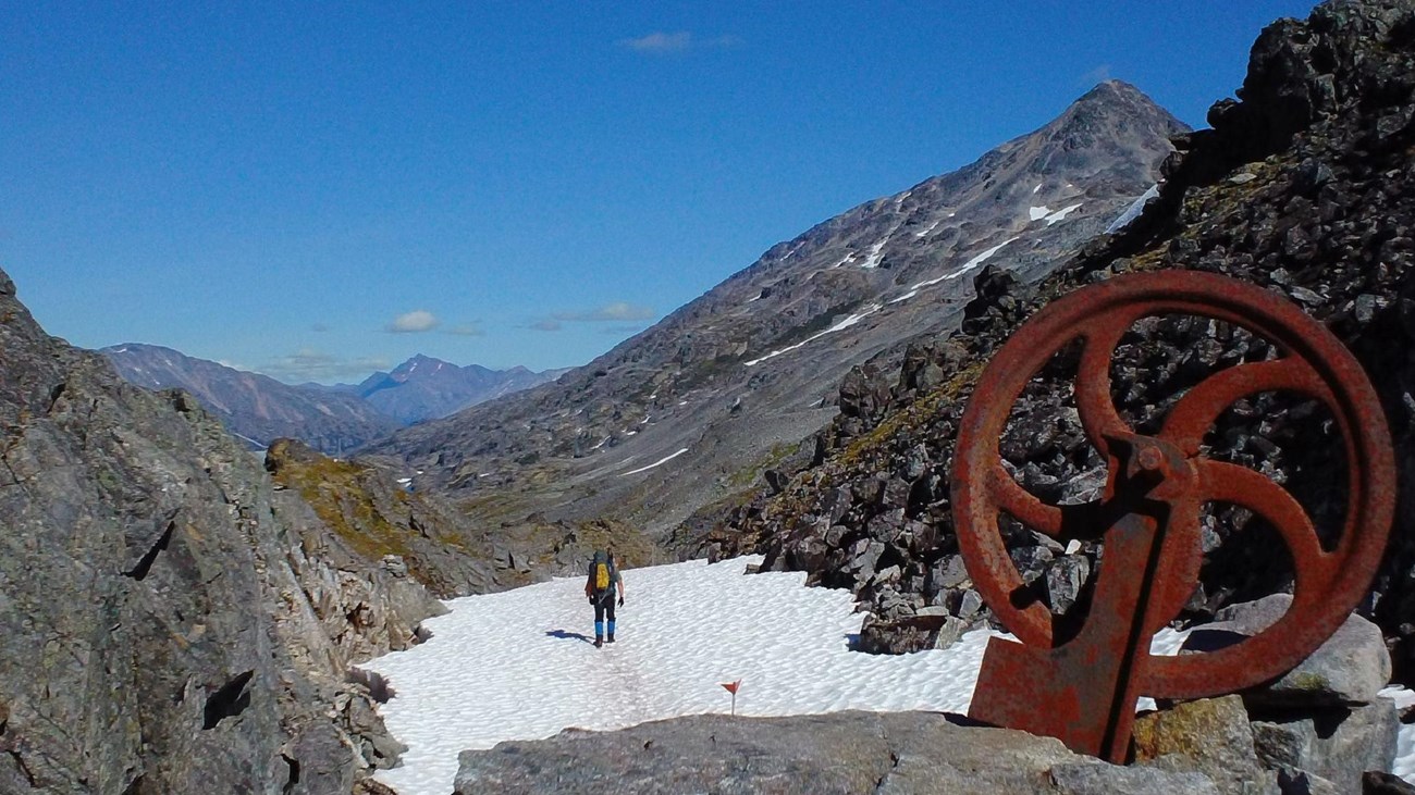 a person on a snow patch between rocky cliffs with a rusted metal wheel in foreground