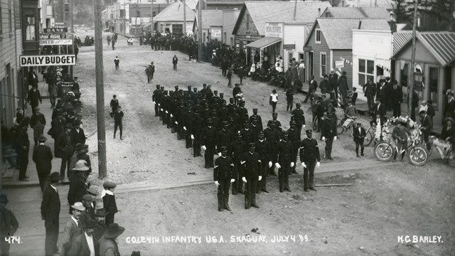 Formation of soldiers standing at attention in a street with onlookers.
