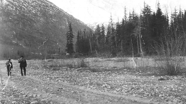 Black and white photo of two people on a dry riverbed with mountains and trees in background