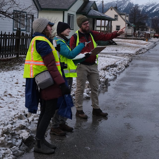 3 people in safety vests stand discussing and looking at buildings.