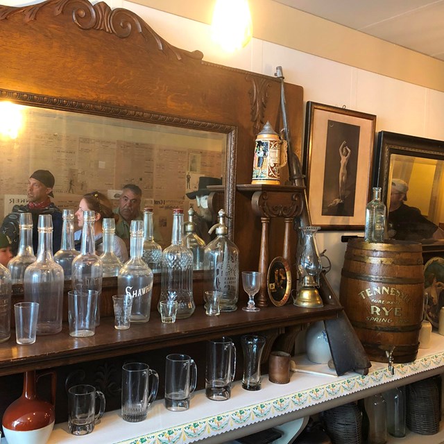 Bottles, glasses, and other items line an old back bar with a mirror behind them