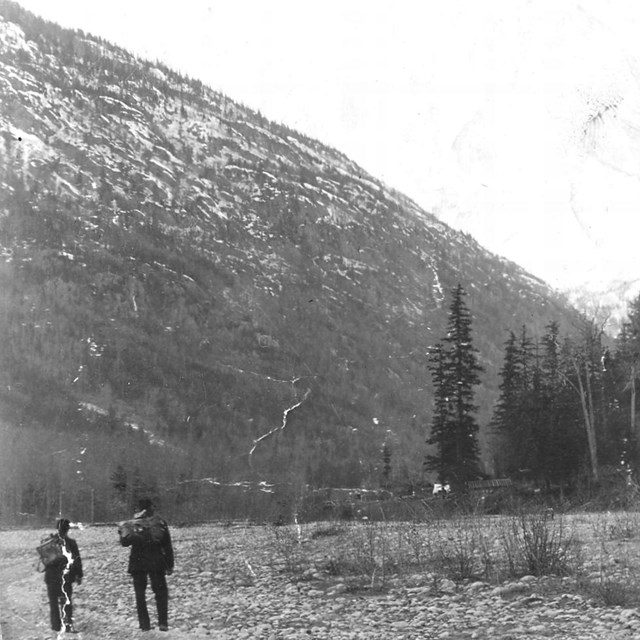 Black and white photo of two people on a dry riverbed with mountains and trees in background