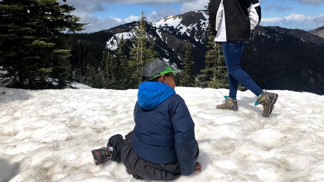 Kid sitting in snow with person walking by.