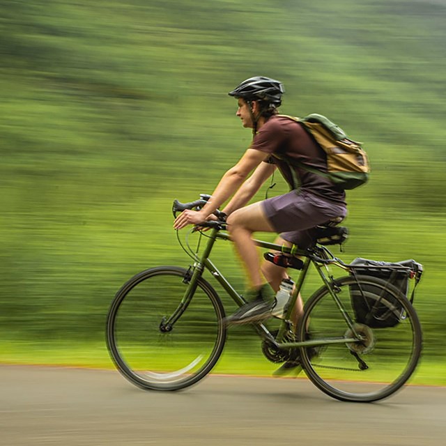Man in brown clothes rides a black bike against a blurry green background implying a fast speed.