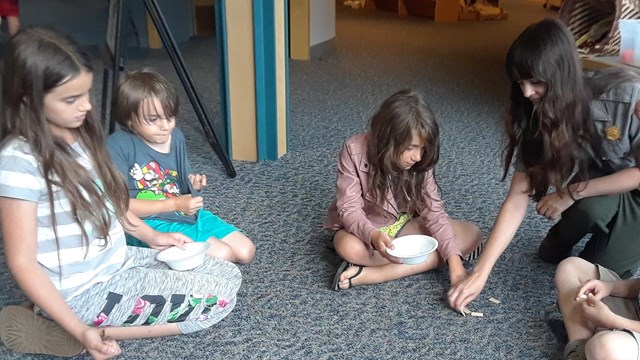 4 kids and female ranger sit in circle on floor playing a game with bowls, sticks, and wooden chips.