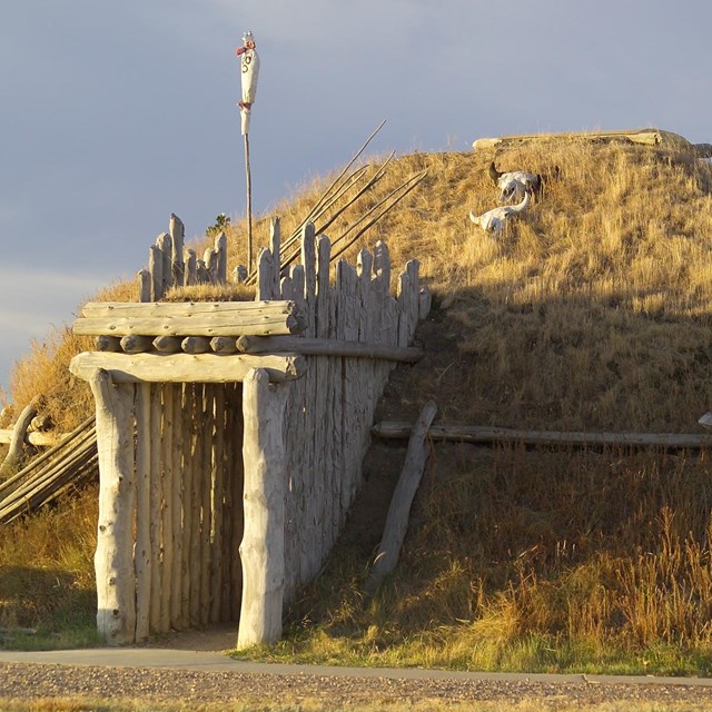 Life-size earthlodge with door facing east. Circular lodge has wooden logs, sod, & dried grasses.