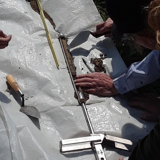 Tarp on grass with soil packed into cylindrical push tube tool. Several hands work with the tool.