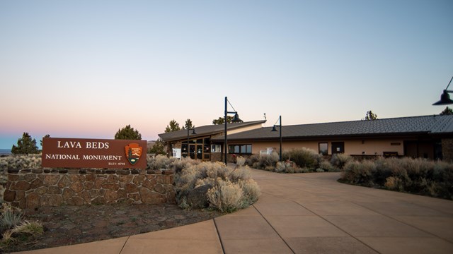 A building with a sign in front reading Lava Beds National Monument at dusk.