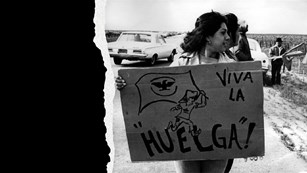 A young woman stands with a sign that reads "Viva la Huelga."