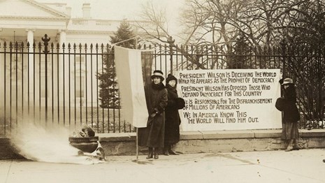 National Woman's Party members outside the White House stand with banenr