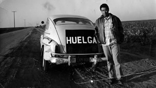 Cesar Chavez standing in front of a car with the word Huelga (strike) on a poster.
