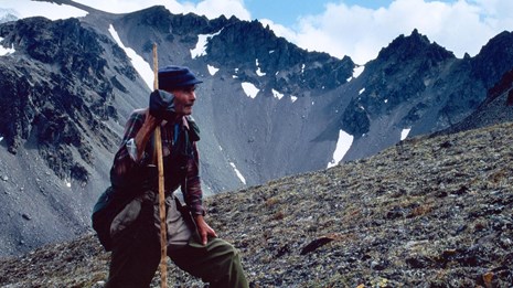 Photo of a man with a tall walking stick standing in alpine tundra with jagged mountains behind him.