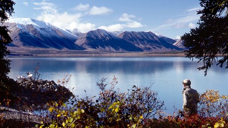 Photo of a man looking away from the camera towards a lake bound by tall mountains.