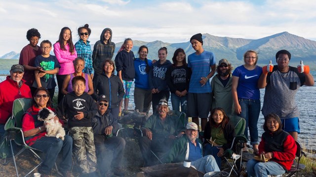 A group of people varying in age stand on shore in front of water and mountains. 