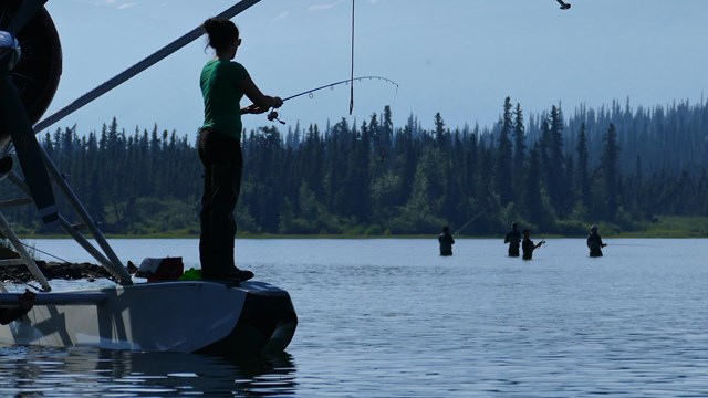 An angler fishes while standing on the float of a plane on a lake with mountains in the background