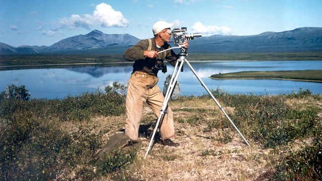 Vintage photo of a man filming with an old movie camera on a tripod near a blue lake and mountains.