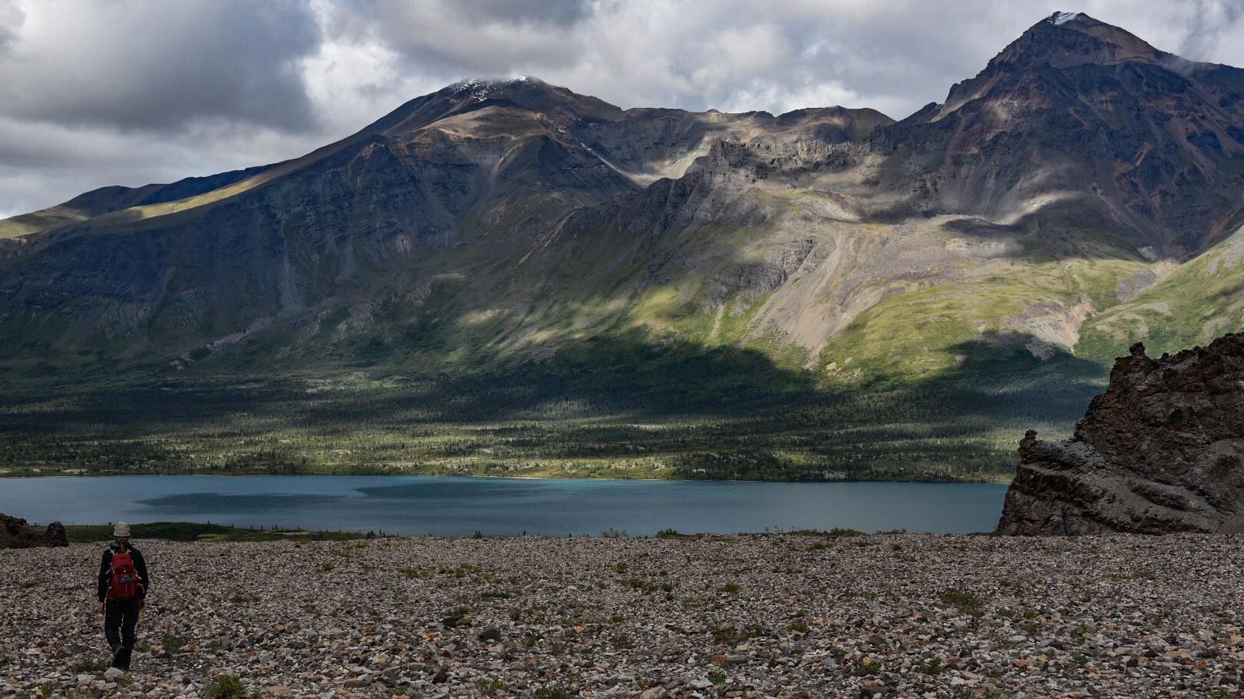 A hiker walks on a rocky riverbed toward a blue lake and dramatically lit mountains