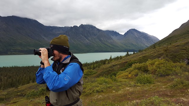 Photo of a park volunteer taking a picture with a blue lake and tall mountains in the background.
