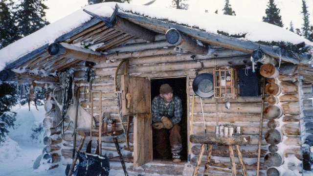 Man wearing blue flannel shirt and brown pants sits in open doorway of a log cabin on a winter day.