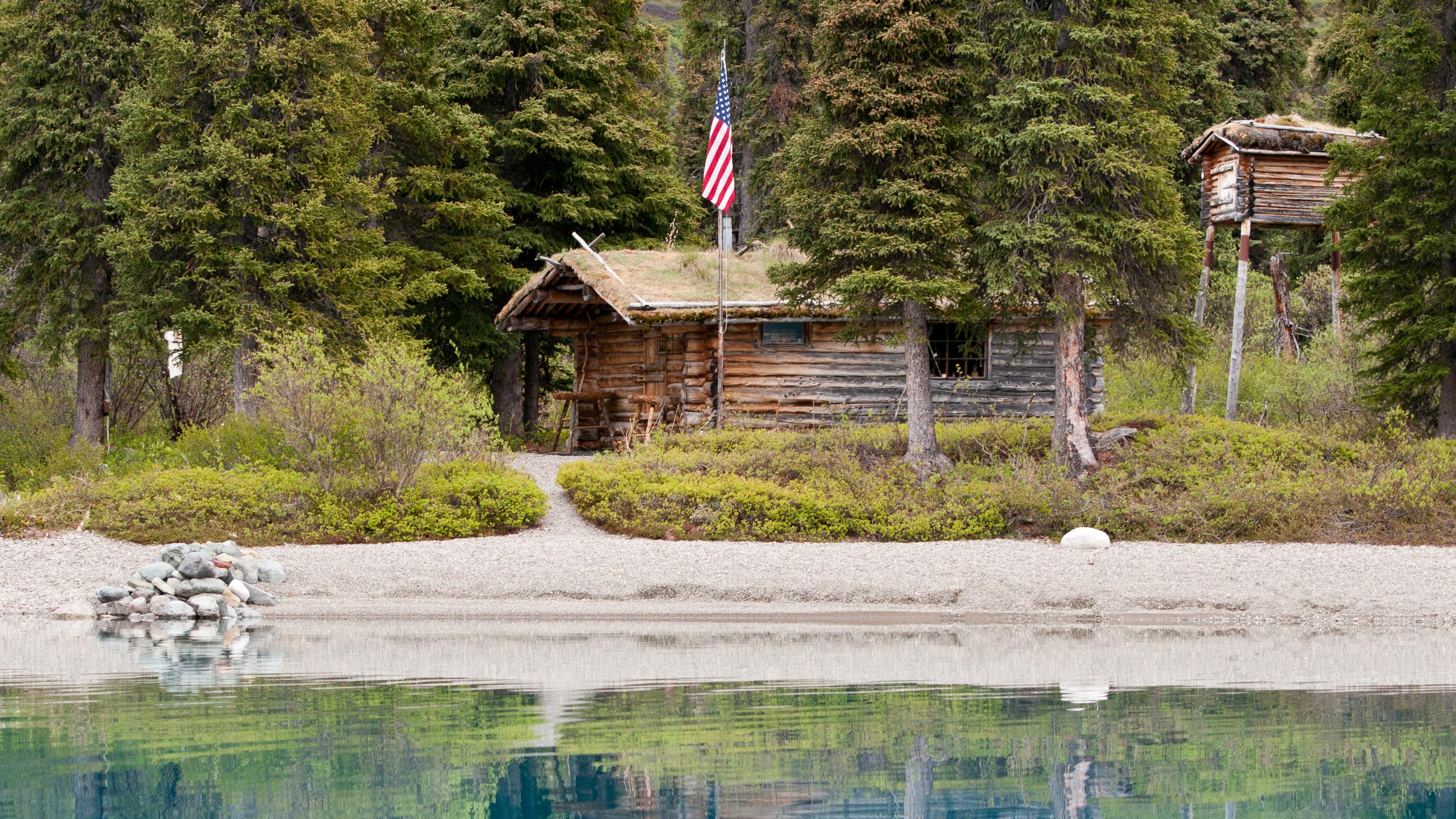 Calm blue water reflects a log cabin with a cache and American Flag surrounded by coniferous trees.